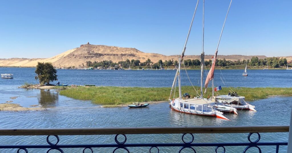 View from a balcony overlooking the Nile River in Aswan, Egypt, with traditional sailboats docked near a lush island and desert hills in the background — a serene scene that captures the allure of 5-star Aswan hotels and helps travelers decide where to stay in Aswan Egypt.
