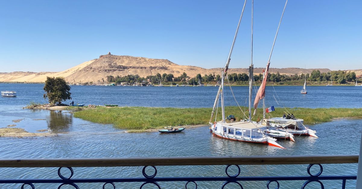 View from a balcony overlooking the Nile River in Aswan, Egypt, with traditional sailboats docked near a lush island and desert hills in the background — a serene scene that captures the allure of 5-star Aswan hotels and helps travelers decide where to stay in Aswan Egypt.