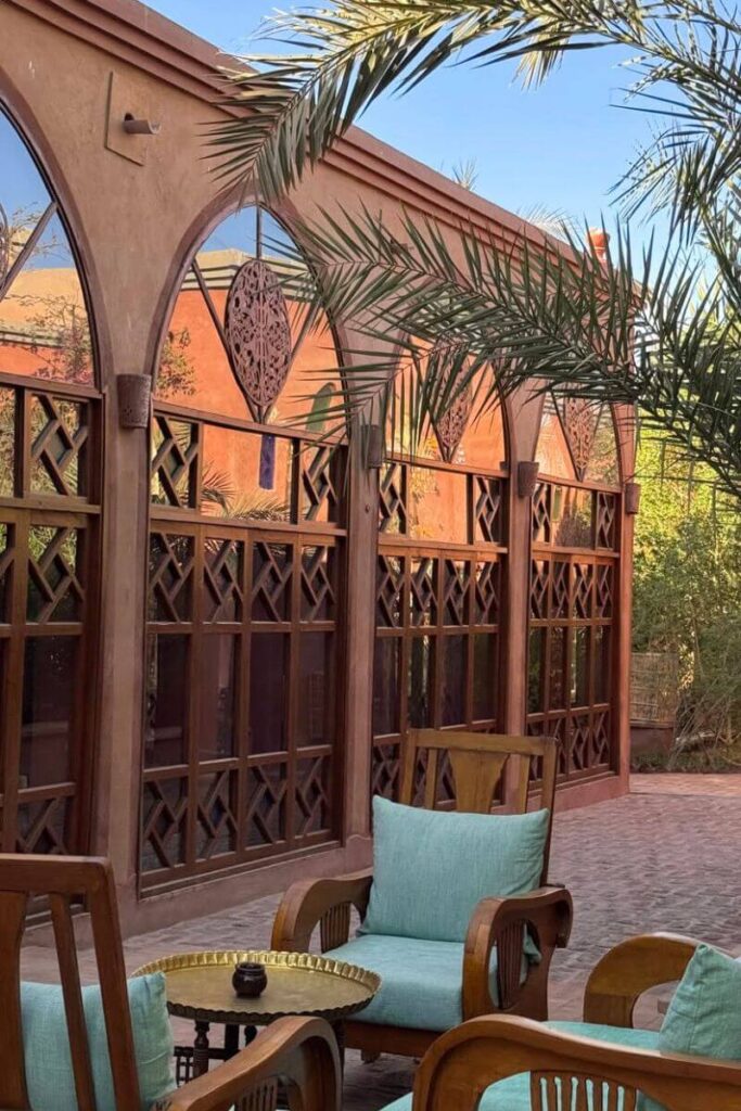 Charming outdoor lounge area at Al Moudira Hotel with carved wooden chairs, mint green cushions, and intricate latticework windows under a palm frond canopy. This intimate setting offers a peaceful retreat, ideal for couples or solo travelers seeking boutique hotel accommodation in Luxor. The architecture blends North African and Mediterranean influences, making it one of the best hotels in Luxor Egypt for design lovers.