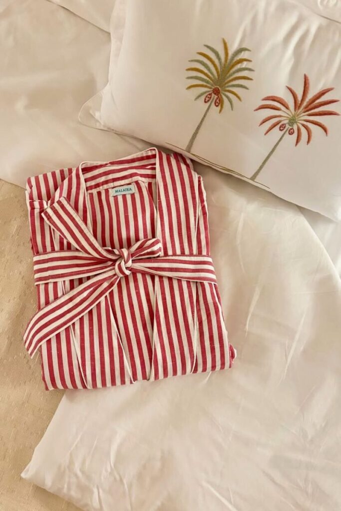 Striped red and white robe labeled “Malaika” neatly folded on a white hotel bed next to a palm tree-embroidered pillow, adding a handmade, luxurious touch. This cozy scene highlights attention to detail at one of Luxor’s boutique hotels. Ideal for guests seeking authentic and thoughtfully designed accommodation in Luxor.