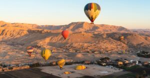 Several colorful hot air balloons lift off over desert hills at sunrise, showcasing the dramatic beauty of hot air ballooning in Luxor Egypt. The golden lighting enhances the visual appeal of this classic Luxor adventure.