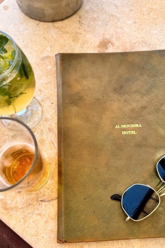 Close-up of a marble table holding a refreshing mojito, a half-full glass of beer, and a leather menu embossed with "Al Moudira Hotel" in gold text, with a pair of black sunglasses beside it. The artisanal setup reflects the character of Al Moudira, one of the most elegant boutique hotels in Luxor. Ideal for travelers searching for unique places to stay in Luxor with charm and sophistication.