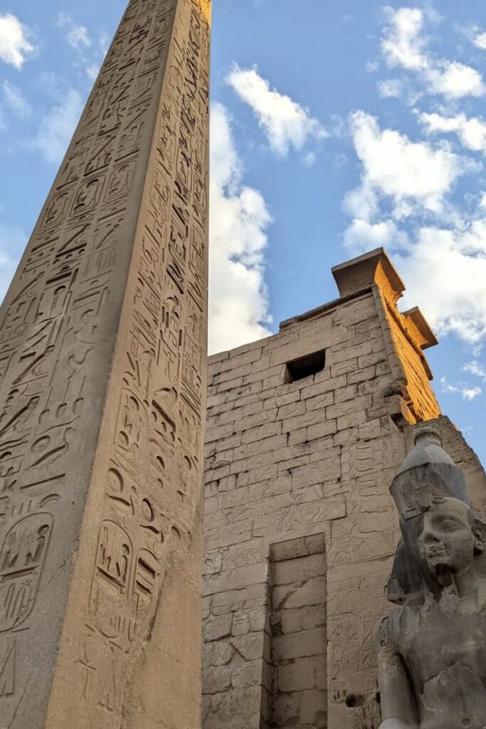 Upward angle of a towering obelisk inscribed with hieroglyphs next to a statue of Ramses II and temple walls, likely from Luxor Temple. A key spot when planning what to do in Luxor, especially for lovers of monumental history.
