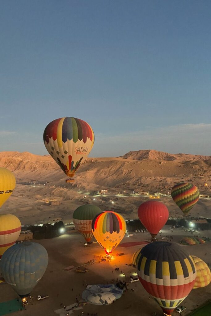 A multicolored balloon labeled “SAFARI BALLOONS” floats gracefully above illuminated desert ground and clustered balloons below, just before sunrise. This vivid depiction of a hot air balloon in Luxor, Egypt captures the pre-dawn anticipation of flight.