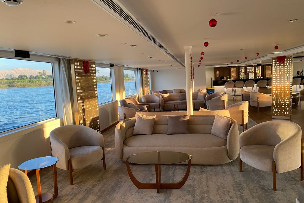 A plush cruise ship lounge with curved beige sofas and red holiday ornaments, overlooking the Nile River. Ideal image for showcasing the comfort and ambiance of an Aswan to Luxor cruise or luxurious Nile cruise experiences.