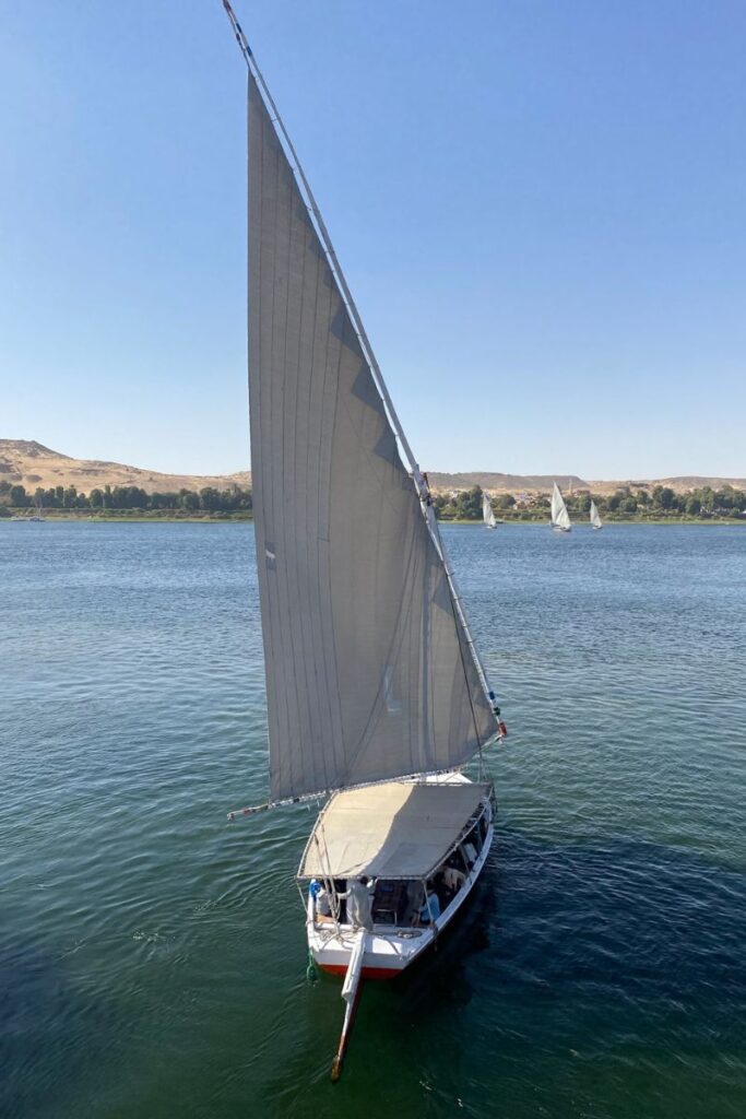 A felucca boat with a towering white sail glides across the Nile River, set against desert hills and calm blue waters. This image complements content on the best Nile cruises, felucca cruises in Aswan, or sailing the Nile.