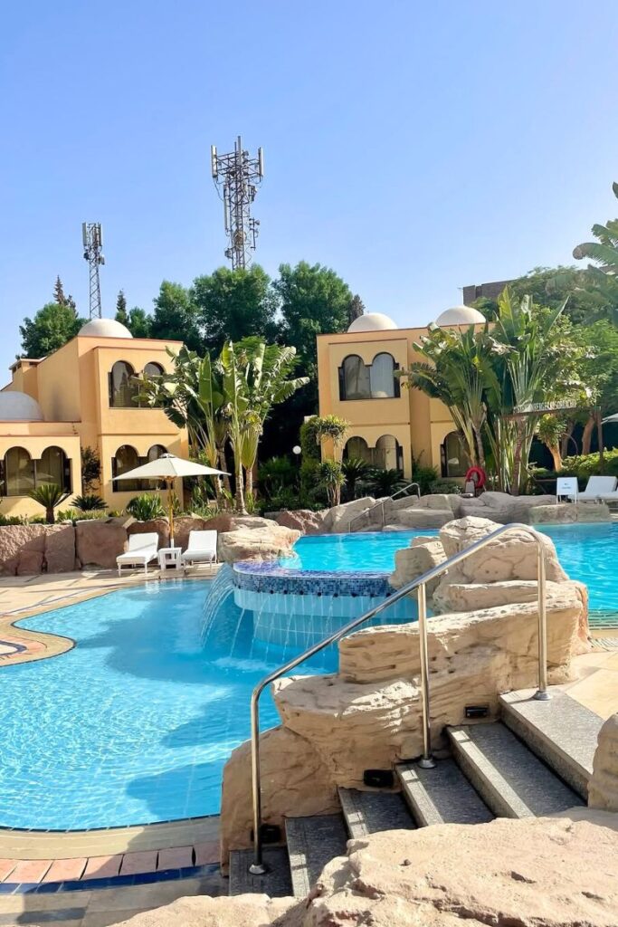 Family-friendly resort pool in Luxor featuring a cascading waterfall, stone detailing, and shallow entry for kids, with banana trees and yellow-domed suites in the background. The inviting space is perfect for families wondering where to stay in Luxor with kids. With resort-style amenities and kid-safe areas, it ranks among the top hotels in Luxor Egypt for family travel.