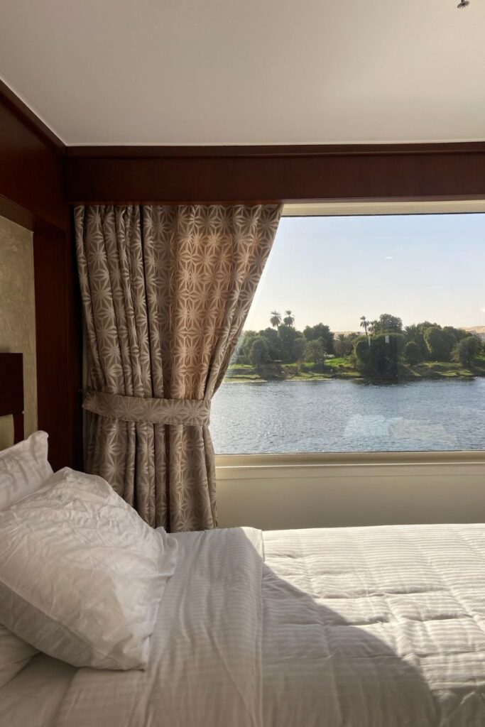 A luxurious bed on a Nile River cruise ship with an expansive window overlooking the Nile and palm trees—perfect for promoting luxury Nile cruises or a romantic cruise from Luxor to Aswan.