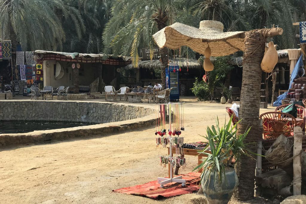 A colorful local market scene at a desert spring in Siwa Oasis, Egypt, with palm trees, handmade jewelry, and a sign reading “Cleopatra Cafe”—offering a taste of things to do in Siwa Oasis and hidden gems in Egypt.