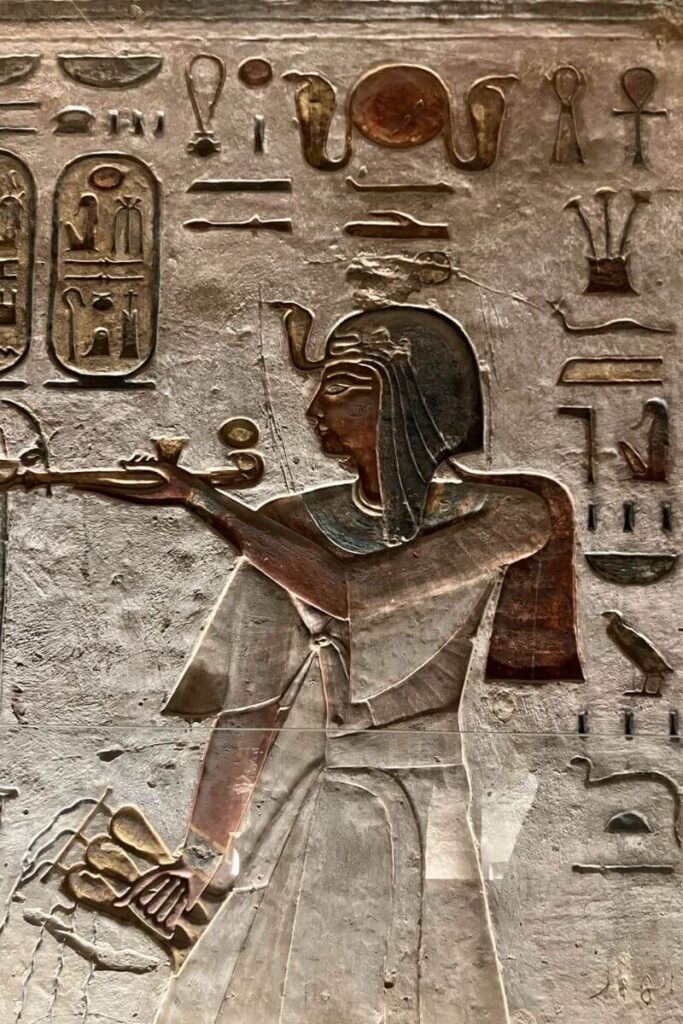 A richly detailed wall relief inside Luxor Valley of the Kings, showcasing a pharaoh wearing a ceremonial headdress and extending his arm to hold symbolic objects. Hieroglyphics and symbolic imagery frame the scene, reflecting the elaborate decorations found within the best tombs in the Valley of Kings Egypt.