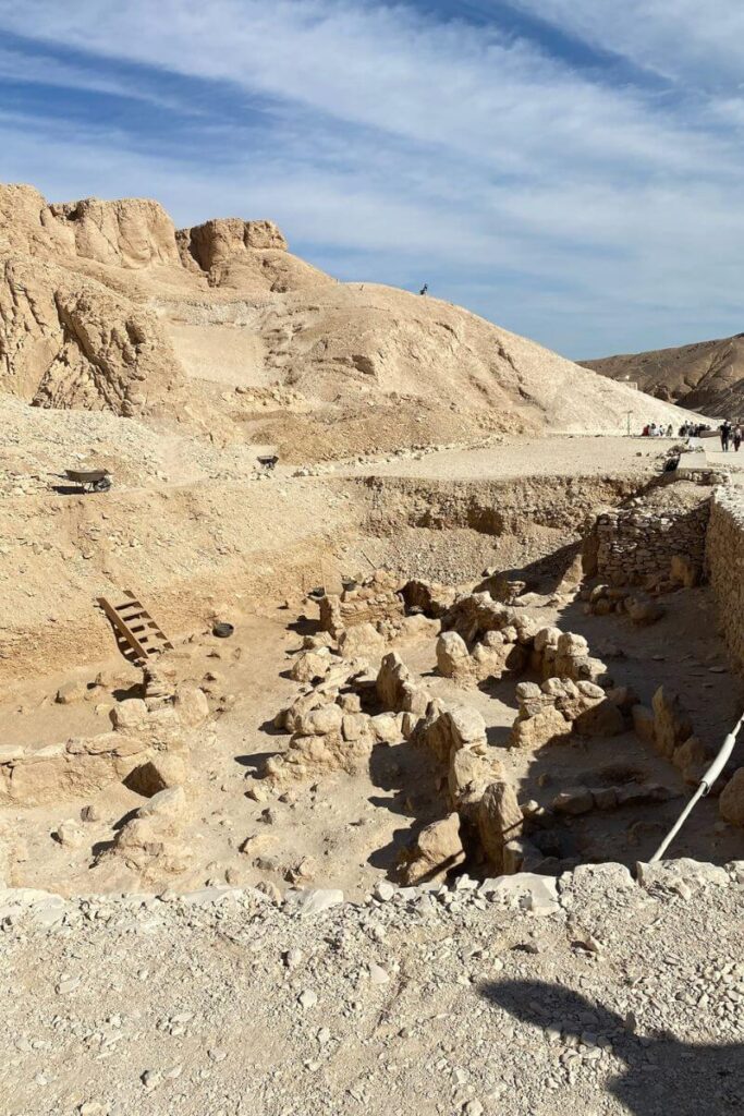 A rocky excavation site in the Valley of the Kings, with uneven stone surfaces and partially exposed ruins. In the distance, visitors can be seen exploring, and a staircase leads down into the dig area, reflecting ongoing archaeological efforts.