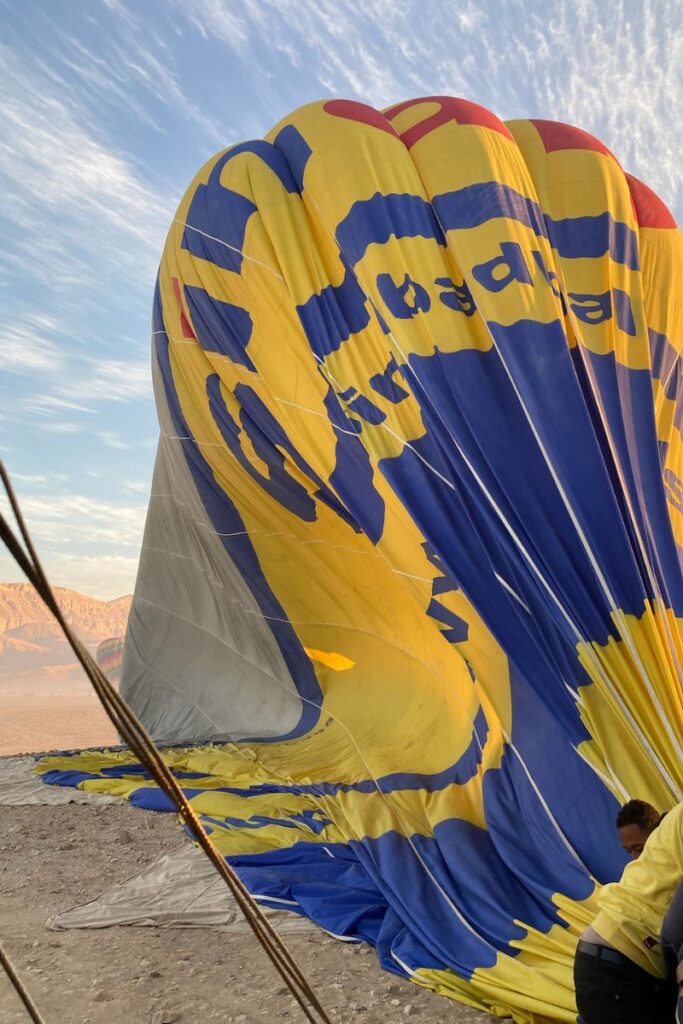 A large deflated yellow and blue hot air balloon lies on the rocky ground in Luxor, with crew members preparing for launch. The text on the balloon reads “Dream Balloon,” offering a look behind the scenes of hot air balloon rides in Egypt.