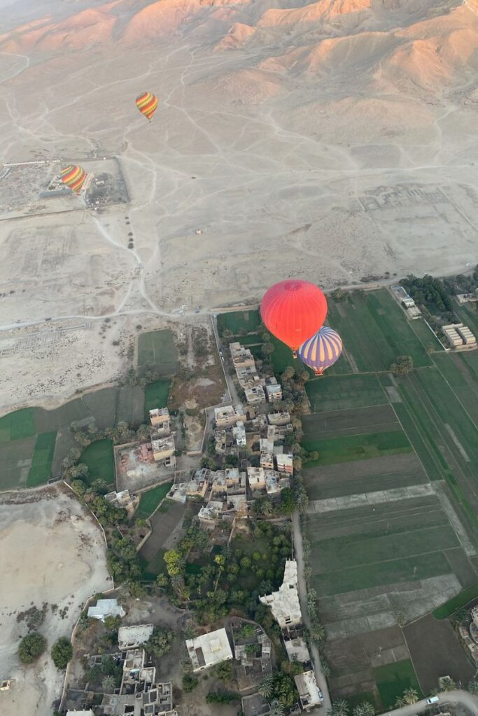 Aerial view of a small village on the edge of green fields and the desert, with vibrant hot air balloons floating overhead. This immersive image highlights the breathtaking contrasts seen during a luxor hot air balloon ride in Egypt