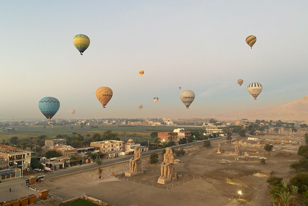 Dozens of colorful hot air balloons drift above the iconic Colossi of Memnon in Luxor, Egypt, during early morning light. The scene highlights the grandeur of a hot air balloon ride Luxor Egypt travelers rave about.