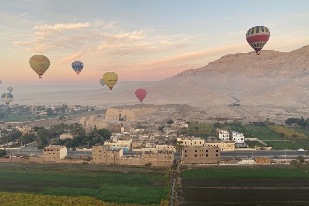 Hot air balloons float above ancient ruins and villages near the mountains of Luxor, Egypt, under a pastel sunrise sky. A breathtaking perspective from one of the most scenic hot air balloon rides in Luxor.