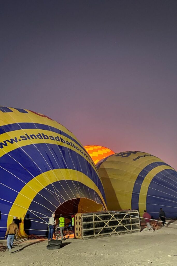 Crew members inflate yellow and blue balloons before sunrise with visible branding for "sindbadballoons.com". This moment captures the preparation stage of hot air ballooning in Luxor, Egypt.