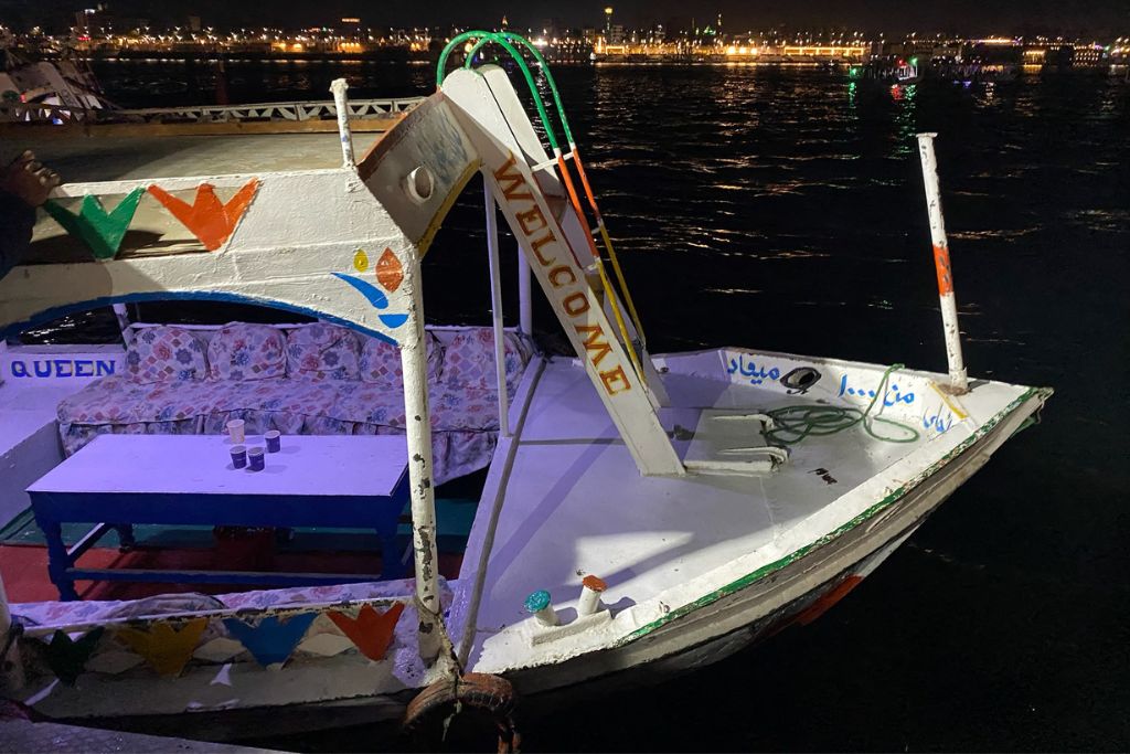 A small, colorfully painted boat labeled "WELCOME" is docked at night with a view of shimmering city lights in the background. While not a balloon scene, this nighttime Nile boat moment adds local flavor to your Luxor adventure.