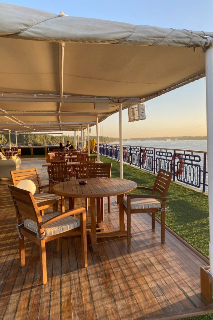 Wooden tables and chairs on a shaded deck of a Nile cruise ship at sunset, with views of the river—great for showcasing the ambiance of a best-rated river cruise or cruise Aswan to Luxor experience.