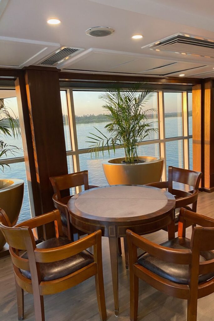Elegant round wooden table with leather chairs by large picture windows overlooking the Nile—an inviting setting aboard a luxury Nile cruise, ideal for highlighting the best Nile cruises Luxor to Aswan.