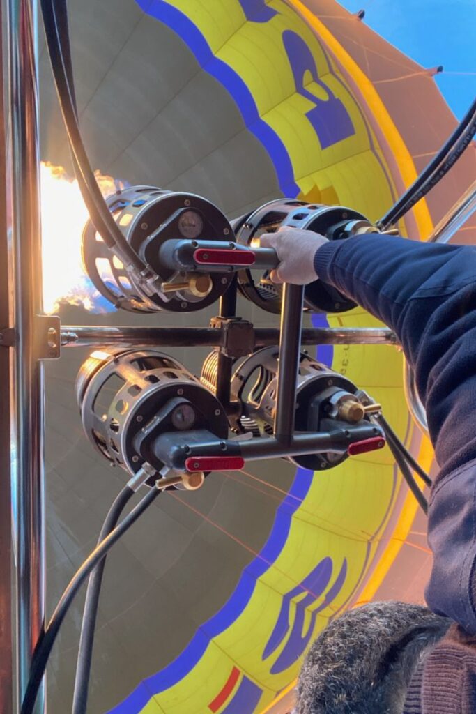 A hand grips the burner controls inside a hot air balloon as bright flames shoot upward, with the interior of a yellow balloon visible above. This action shot offers a close-up of how a Luxor balloon ride is powered.