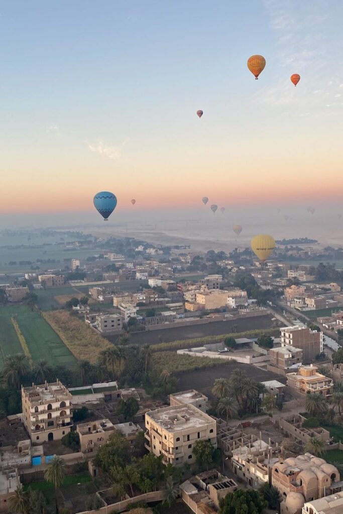 Hot air balloons rise over early morning rooftops and green fields in Luxor, Egypt, as the sun begins to warm the sky. A serene scene that shows why luxor balloons are among the top attractions in Egypt.