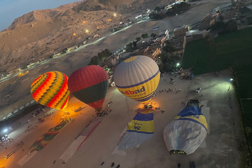 Hot air balloons are preparing for launch at dawn, their flames glowing orange as people gather around. One balloon reads "Sindbad," a popular operator for a Luxor Egypt hot air balloon experience.