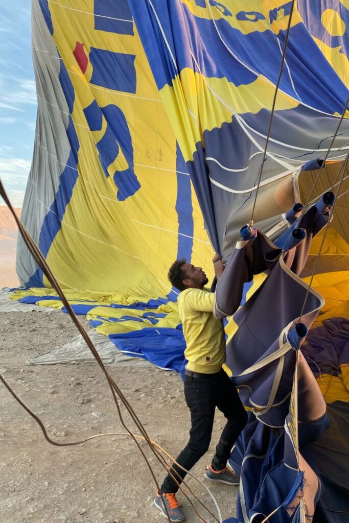 A crew member in a yellow hoodie holds onto the ropes of a large yellow and blue balloon being inflated in the desert. This image illustrates the human effort behind every Luxor hot air balloon ride.