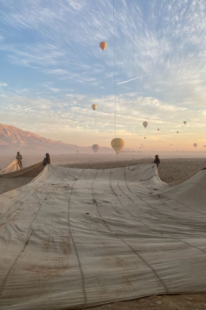 Crew members work with a deflated balloon envelope as hot air balloons take flight under pastel skies in Luxor. This behind-the-scenes look captures the prep phase of a Luxor balloon ride.