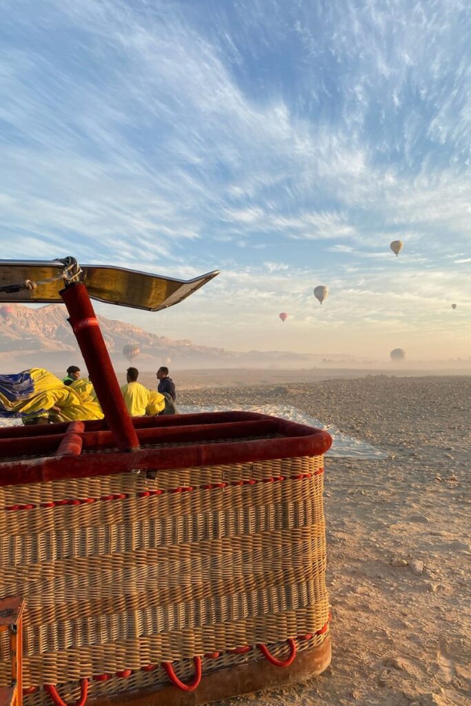 A woven balloon basket with red trim sits on the desert floor while crew members in yellow jackets work in the background. Hot air balloons float in the distance, ready for another magical Luxor Egypt hot air balloon morning.