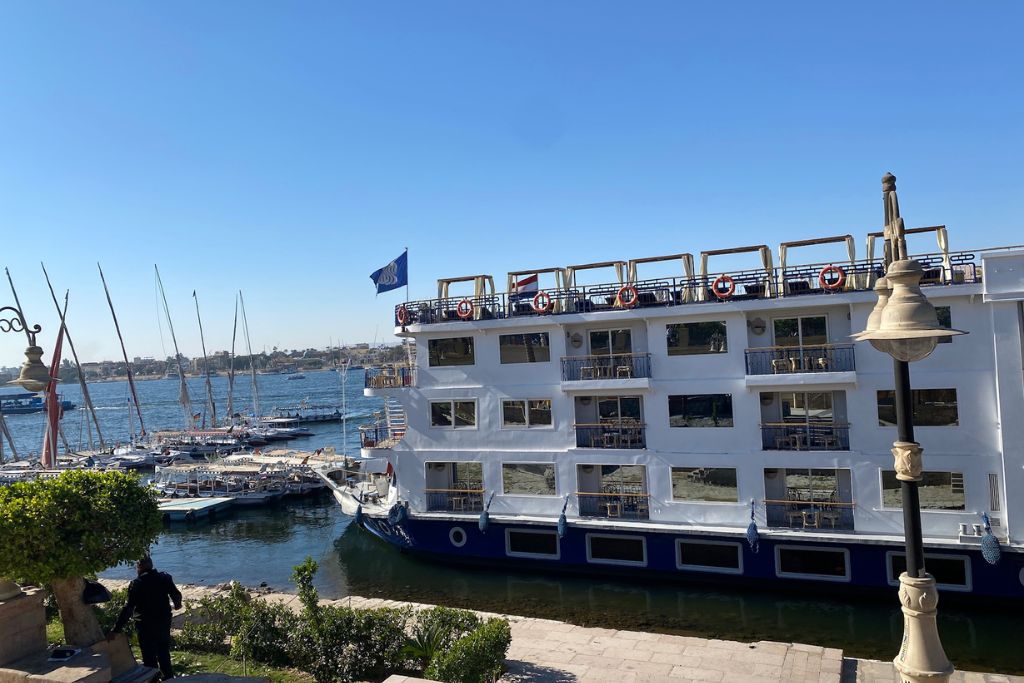 Exterior view of a multi-level Nile cruise ship docked beside felucca boats on the Nile. Great for showcasing the diversity of Nile River cruise ships, from traditional to luxurious options like dahabiya Nile cruises.