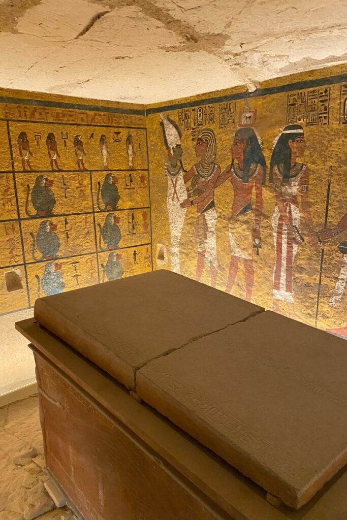 Luxor Valley of the Kings tombs interior chamber with a large stone sarcophagus in the foreground, surrounded by vibrant wall murals depicting Egyptian deities, ritual scenes, and symbols. The yellow and gold tones of the artwork contrast with the worn stone ceiling, emphasizing the historic significance of the burial chamber.