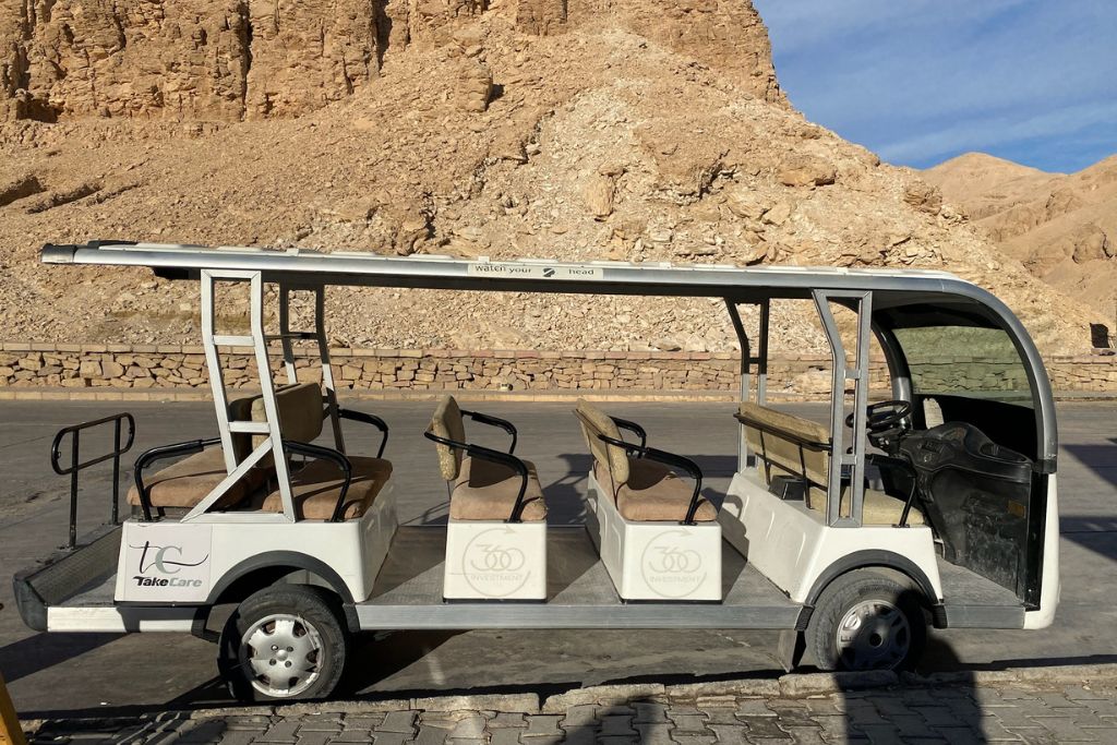 An open-top electric vehicle parked near the base of the cliffs at Luxor Valley of the Kings, with seating for multiple passengers, used to transport visitors around the valley of kings Egypt during tours of the tombs. The rocky landscape in the background highlights the desert terrain surrounding the ancient tombs.