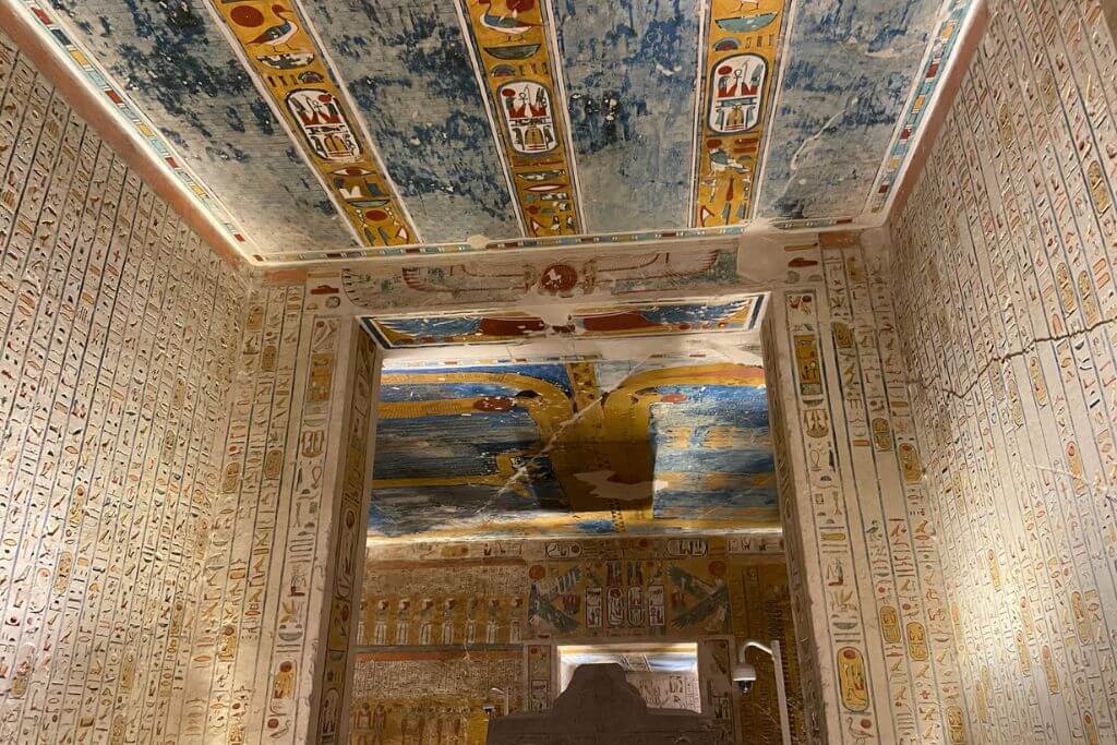 Luxor Valley of the Kings tombs corridor with extensive hieroglyphic text covering the walls, interspersed with images of gods, humans, and mythical creatures. The painted ceiling, in shades of blue and gold, reflects the ancient Egyptians’ emphasis on the heavens in burial traditions.