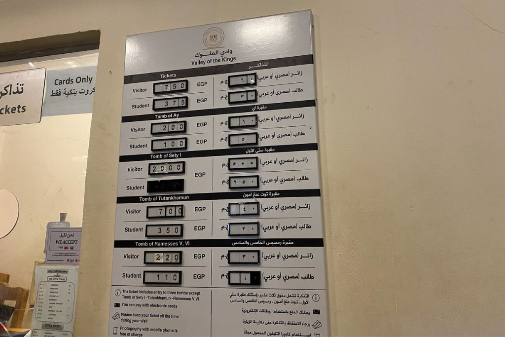 A large white board mounted on a wall, displaying ticket prices in Egyptian pounds for various tombs inside the Valley of the Kings. The board shows separate prices for visitors and students, including popular tombs such as Tutankhamun’s and Seti I’s. An adjacent glass window indicates the location of a ticket counter.