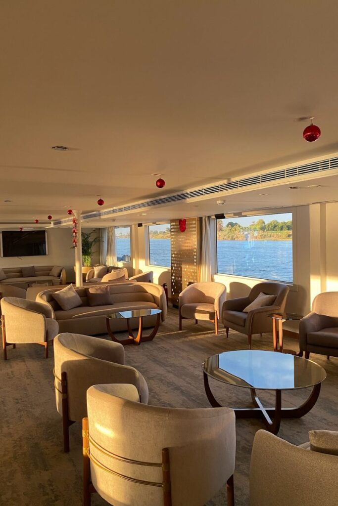 Spacious cruise ship lounge with beige armchairs and wide river-view windows, decorated with red holiday ornaments—highlighting comfort aboard a luxurious Nile cruise or dinner cruise Nile experience.