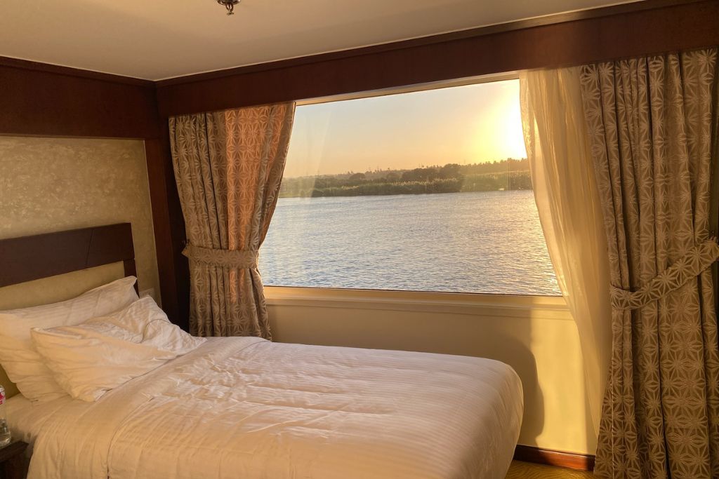 Cozy cruise cabin with a large picture window showing the Nile at sunset, golden light filling the room—fits content about luxurious Nile cruise cabins and romantic river Nile dinner cruise stays.
