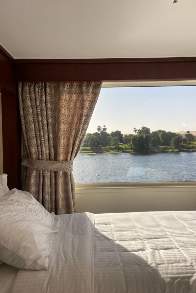 Cozy Nile River cruise cabin with a large window view of the lush riverbank. Ideal for a relaxing stay while exploring places to visit in Aswan, and a peaceful moment on your Aswan itinerary.