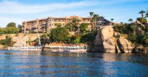 View of the luxurious Old Cataract Hotel perched on the edge of the Nile River, with traditional white-sailed feluccas floating below. This iconic colonial-era hotel is one of the most elegant places to visit in Aswan and a scenic highlight for travelers looking for unique things to do in Aswan, Egypt.