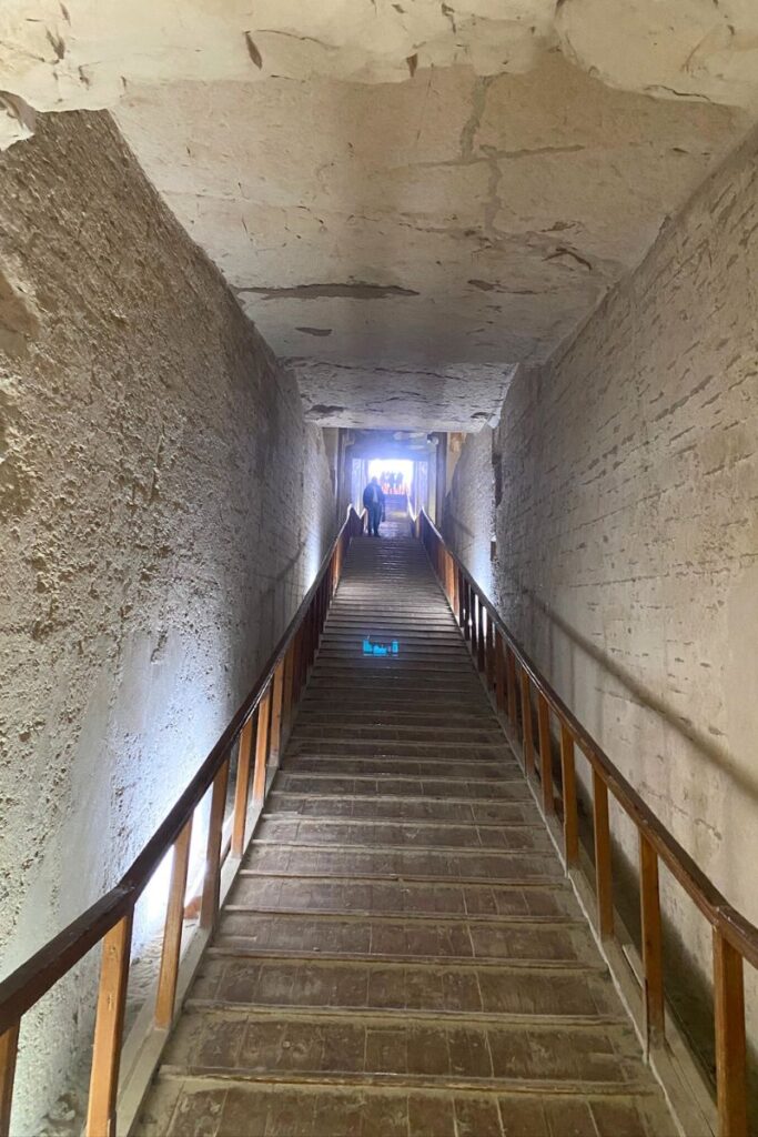 The corridor of Luxor Valley of the Kings tombs shows a long, steep staircase with wooden railings on both sides, bordered by high walls of natural stone. The rough, textured ceiling and sidewalls reveal layers of carved rock, and visitors can be seen silhouetted against the light at the distant exit.