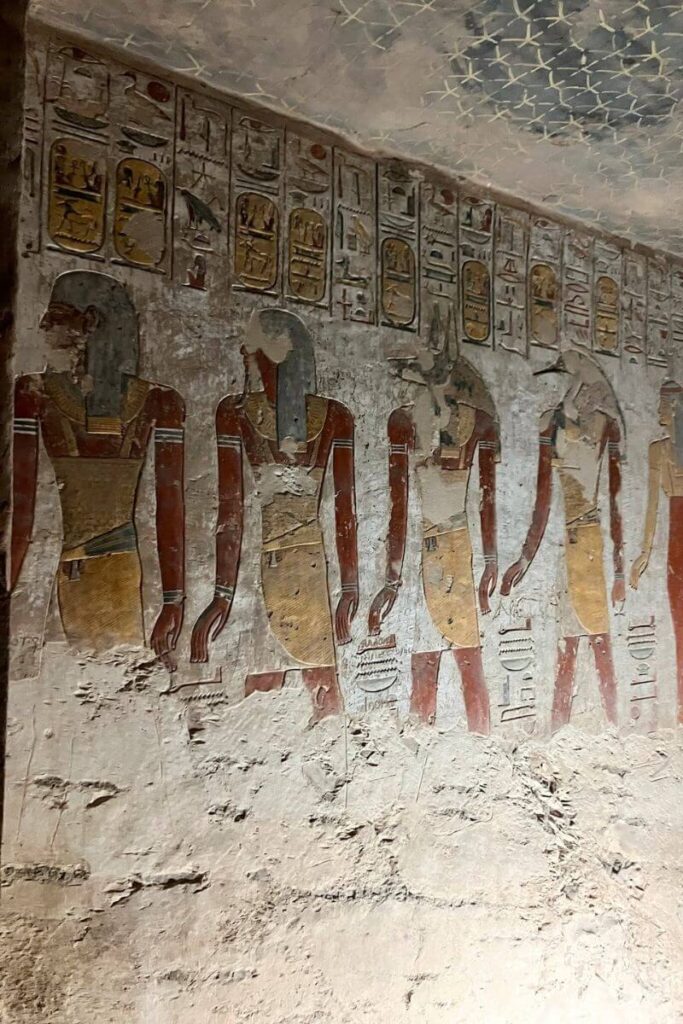 Inside Luxor Valley of the Kings tombs, an ancient wall painting depicts a row of standing Egyptian figures in traditional garb, with detailed accessories and headdresses. Hieroglyphs fill the upper portion of the wall, while the lower part shows weathered stone.