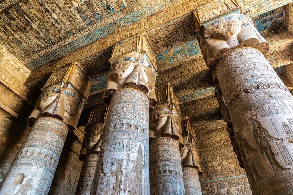 Massive temple columns topped with Hathor-faced capitals under a richly decorated blue ceiling inside the Temple of Hathor at Dendera. One of the most visually stunning places to see in Luxor and a unique attraction near the city.
