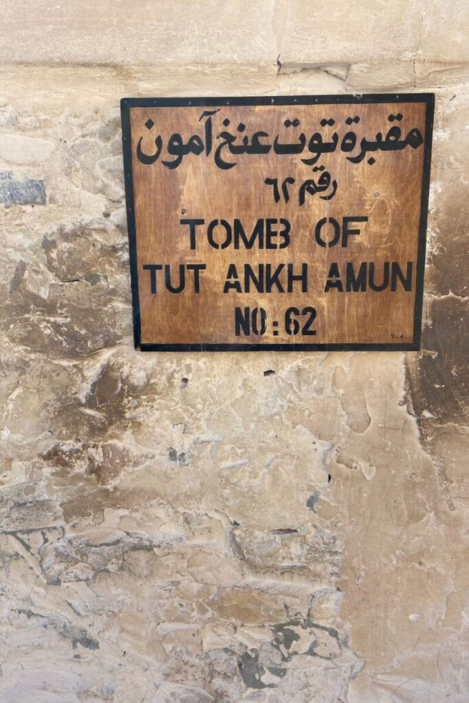The images depict the entrance to the Tomb of Tutankhamun in Luxor Valley of the Kings tombs, marked by a wooden sign with bold black letters in both Arabic and English, indicating "Tomb of Tutankhamun No:62." The sign is mounted on a rough stone wall, showcasing the weathered texture of the valley’s rock face. The surroundings feature light beige and grey tones, with the entrance seamlessly blending into the natural landscape of the Valley of the Kings in Egypt.