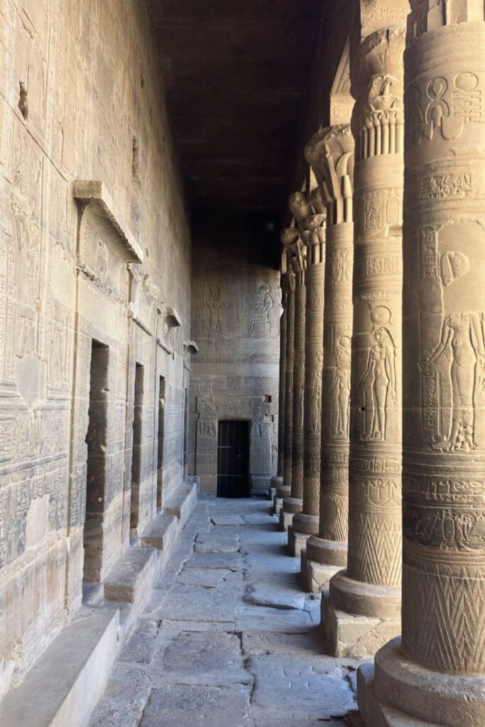 View down a narrow, column-lined corridor of an ancient Egyptian temple covered in carvings and hieroglyphs. A stunning stop for history lovers exploring what to do in Aswan and top attractions in Aswan.