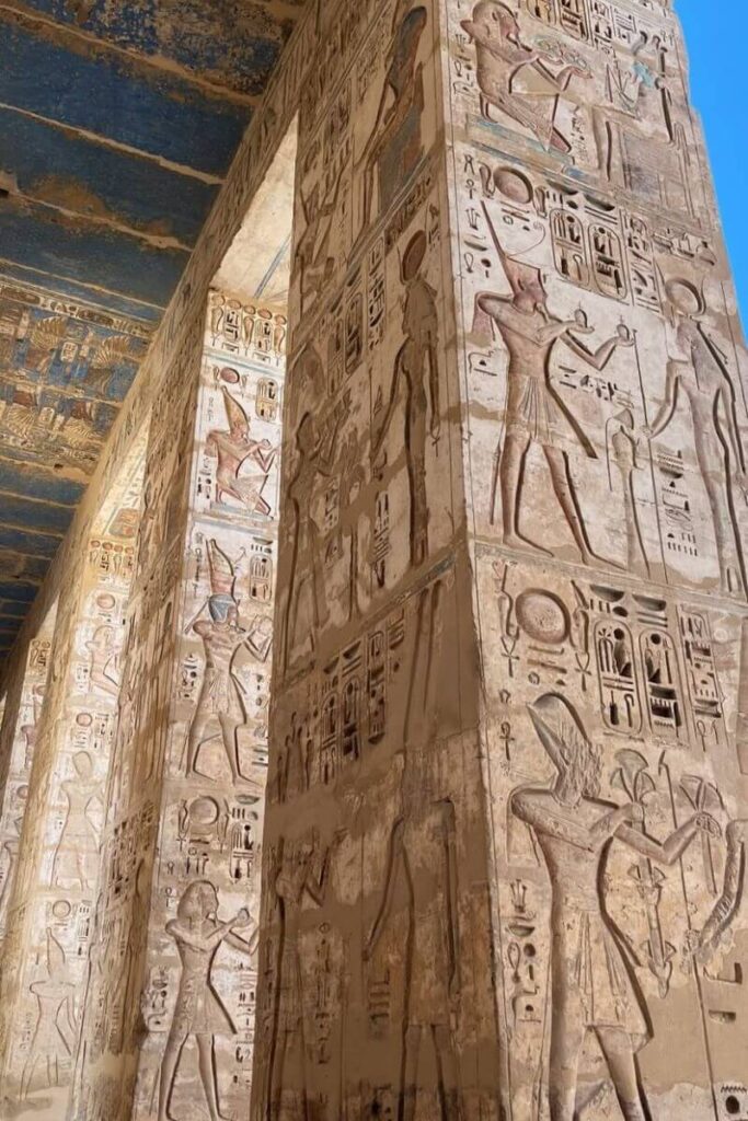 Tall columns covered in carved and painted reliefs showing ancient Egyptian pharaohs and gods, with remnants of a blue ceiling above. A classic example of the intricate temples and places to see in Luxor for history lovers.