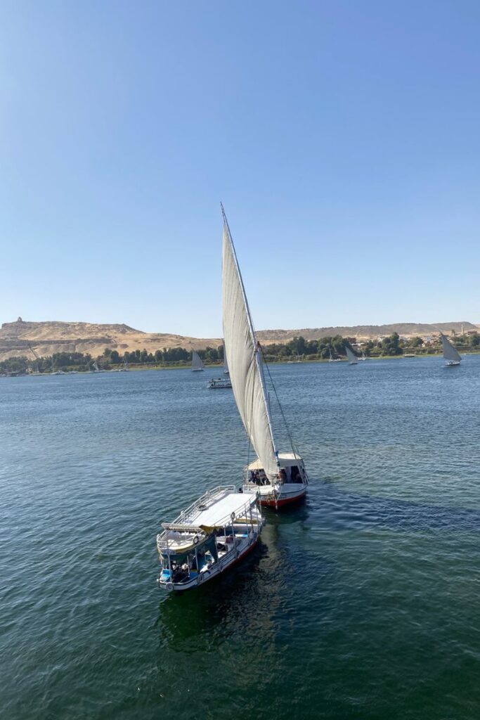 Two traditional sailboats glide across the Nile River under clear blue skies with desert hills in the background. A classic scene representing what to see in Aswan on a Nile cruise or felucca ride.