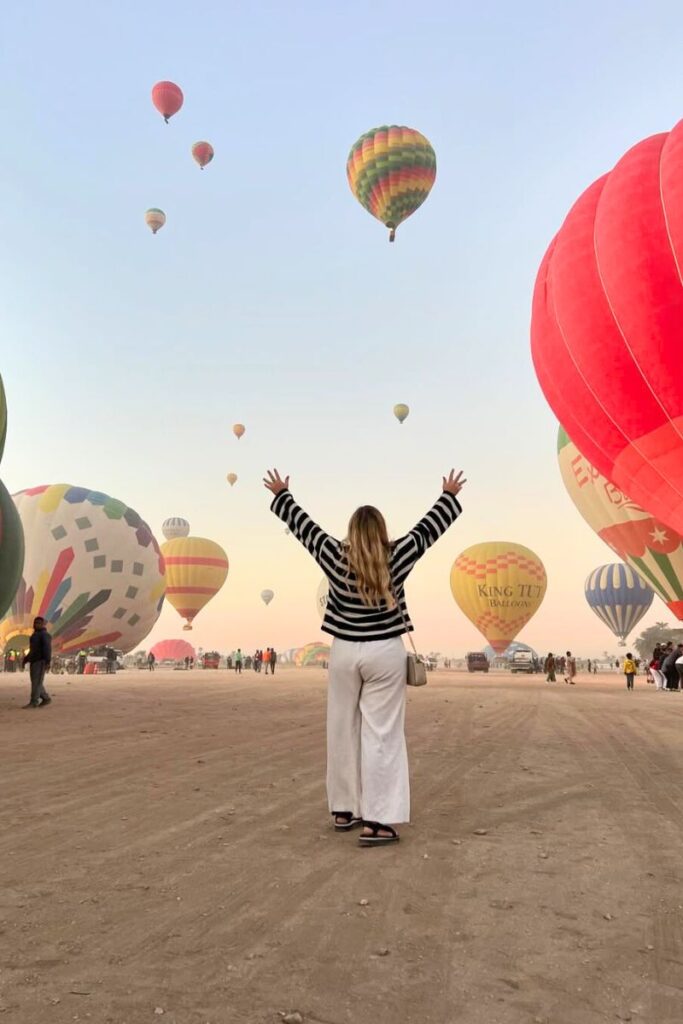 Dozens of vibrant hot air balloons inflate and lift off at dawn in Luxor as a woman stands with arms raised in awe. Luxor hot air balloons are one of the most iconic experiences in Egypt.