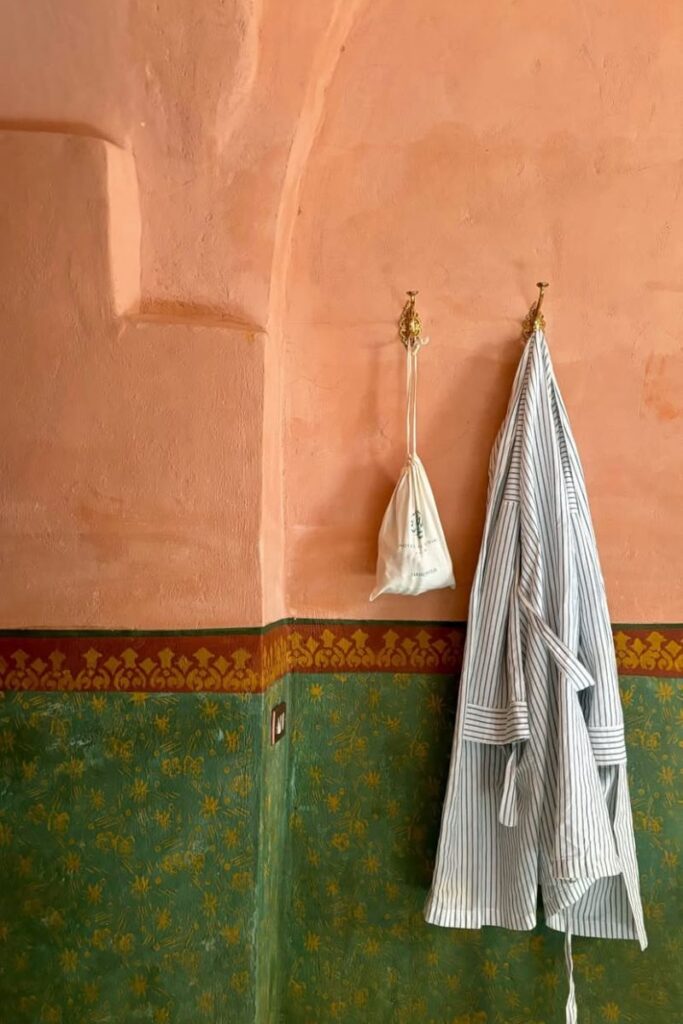 Soft robe and a linen bag hanging on brass hooks inside a warmly painted room with green and red wall detailing. Stylish comfort after experiencing hot air balloons in Luxor at sunrise.