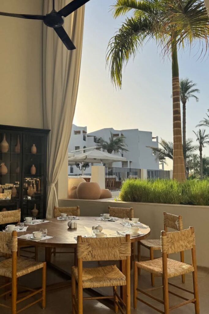 A round wooden breakfast table with rustic chairs is set on a shaded patio, looking out at palm trees and white resort buildings. Captures the atmosphere of the best hotels in Hurghada for couples and the best sunrise hotel in Hurghada.