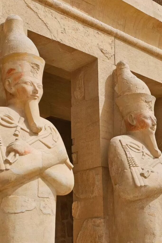 Two statues of pharaohs standing guard at a temple entrance, arms crossed and adorned with royal headgear, highlighting classic Egyptian monuments and temples in Egypt.