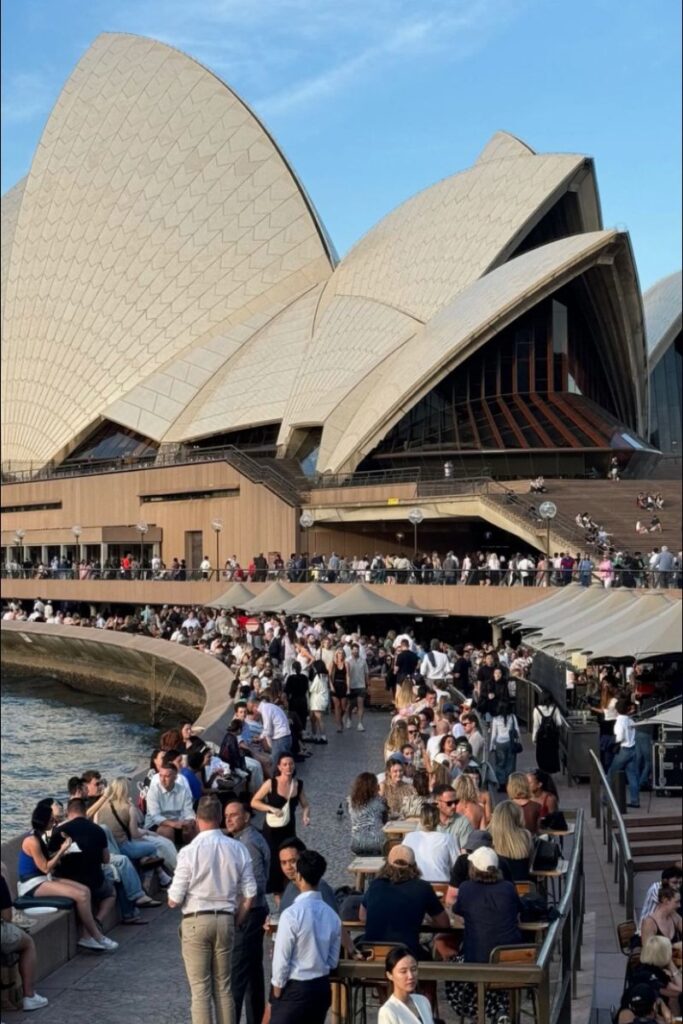 A crowded waterfront promenade at the Sydney Opera House with people dining and socializing under umbrellas. A great fit for posts about solo female travel Australia, best destinations for solo female travelers, or traveling alone in Australia.